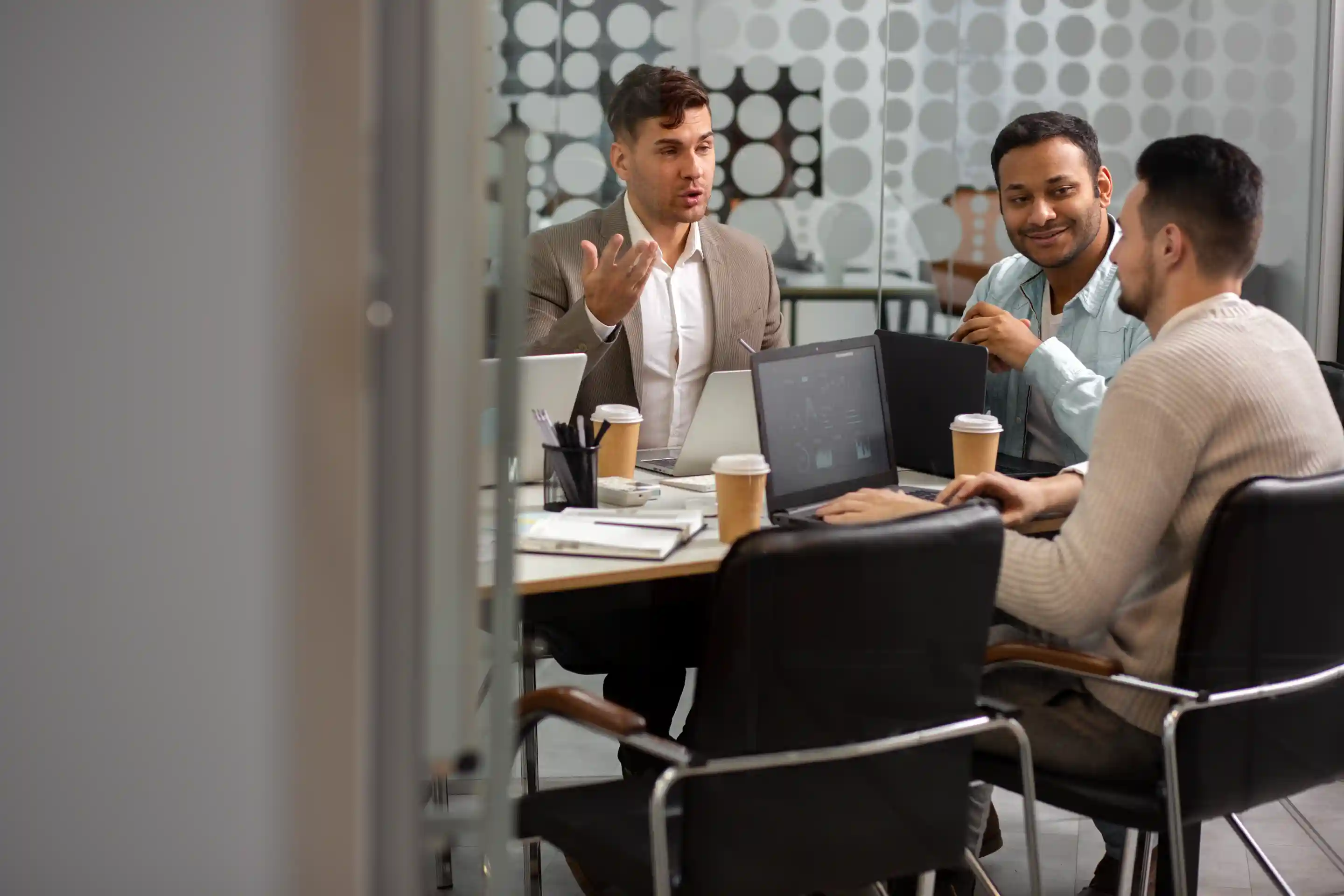 Three professionals in a business meeting, reviewing data on laptops with coffee cups on the table