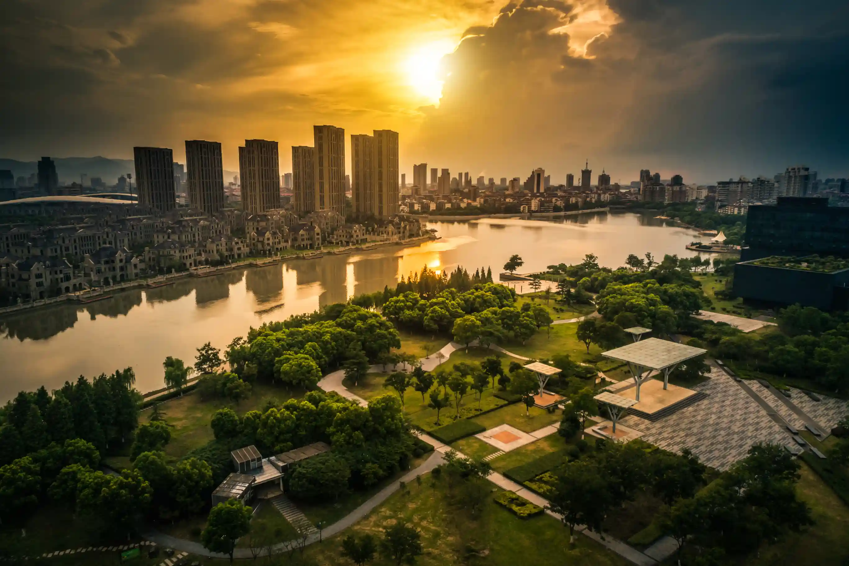 Golden-hour aerial view of a city skyline with a reflective river, green park, and residential towers